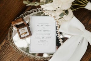 Wedding invitation on a mirrored tray along with wedding rings and a pearl necklace